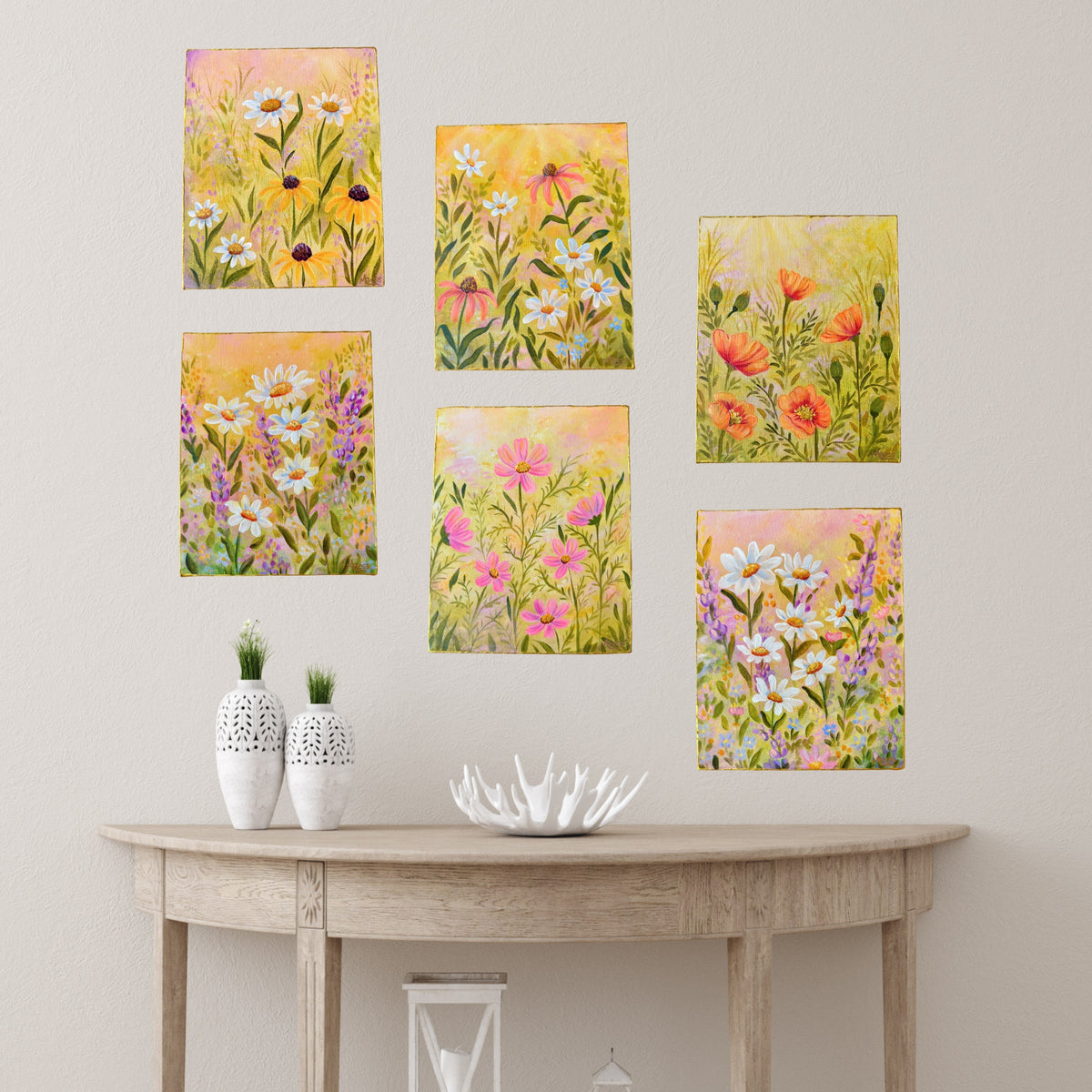 Six vertical floral art prints with colorful wildflower designs are arranged on a light beige wall above a curved light wood console table with white decorative vases and a white bowl.