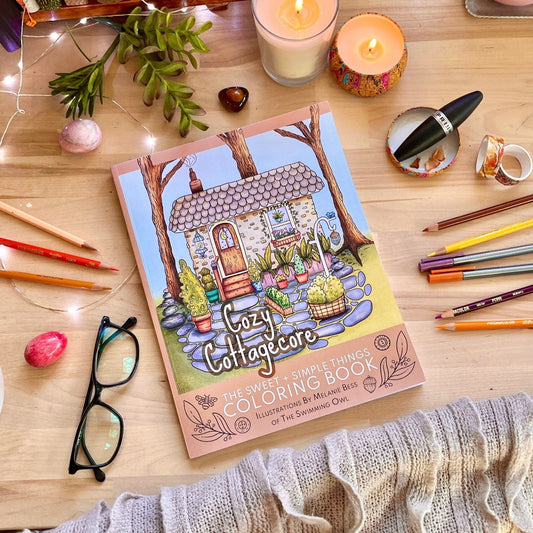 Coloring book titled 'Cozy Cottagecore' on a wooden table with coloring supplies and a cup of coffee.