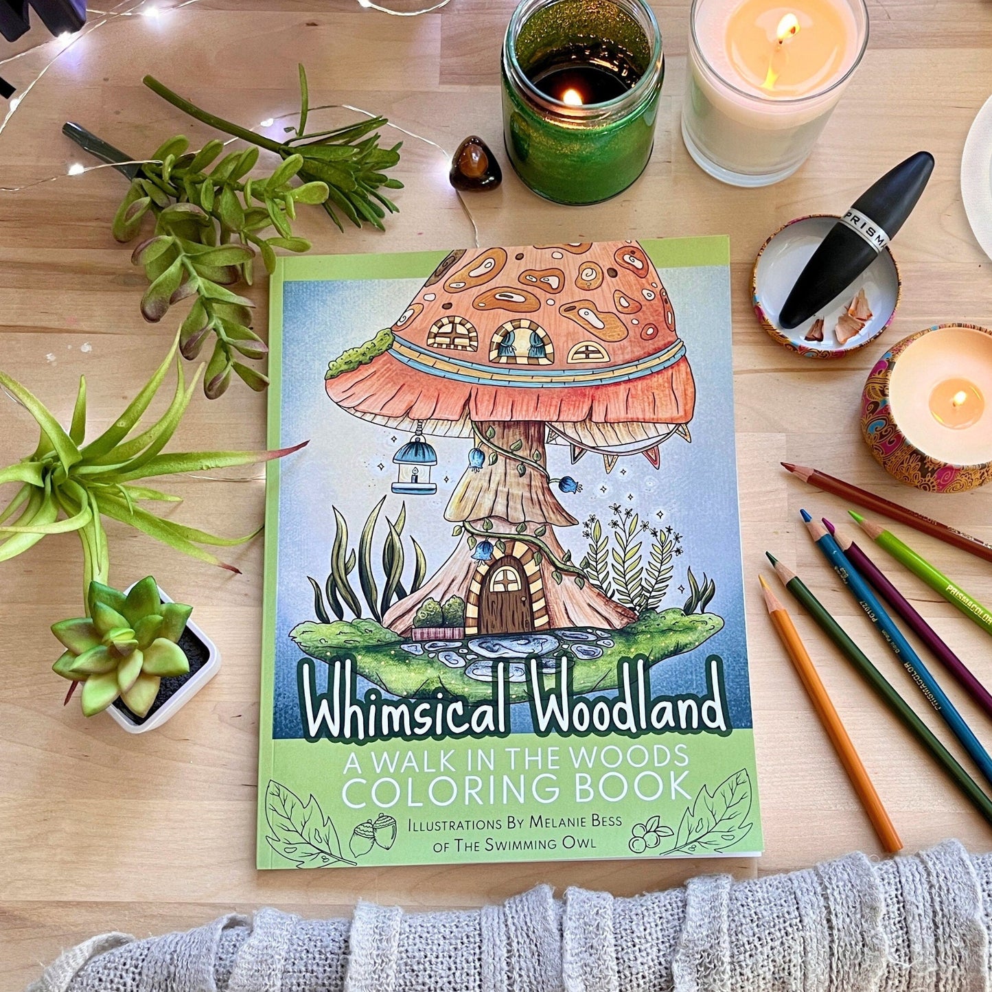 Coloring book titled 'Whimsical Woodland' on a table with candles, plants, and coloring pencils.